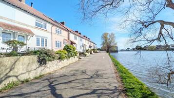 A paved walkway alongside a river, with houses on one side and a grassy embankment on the other.