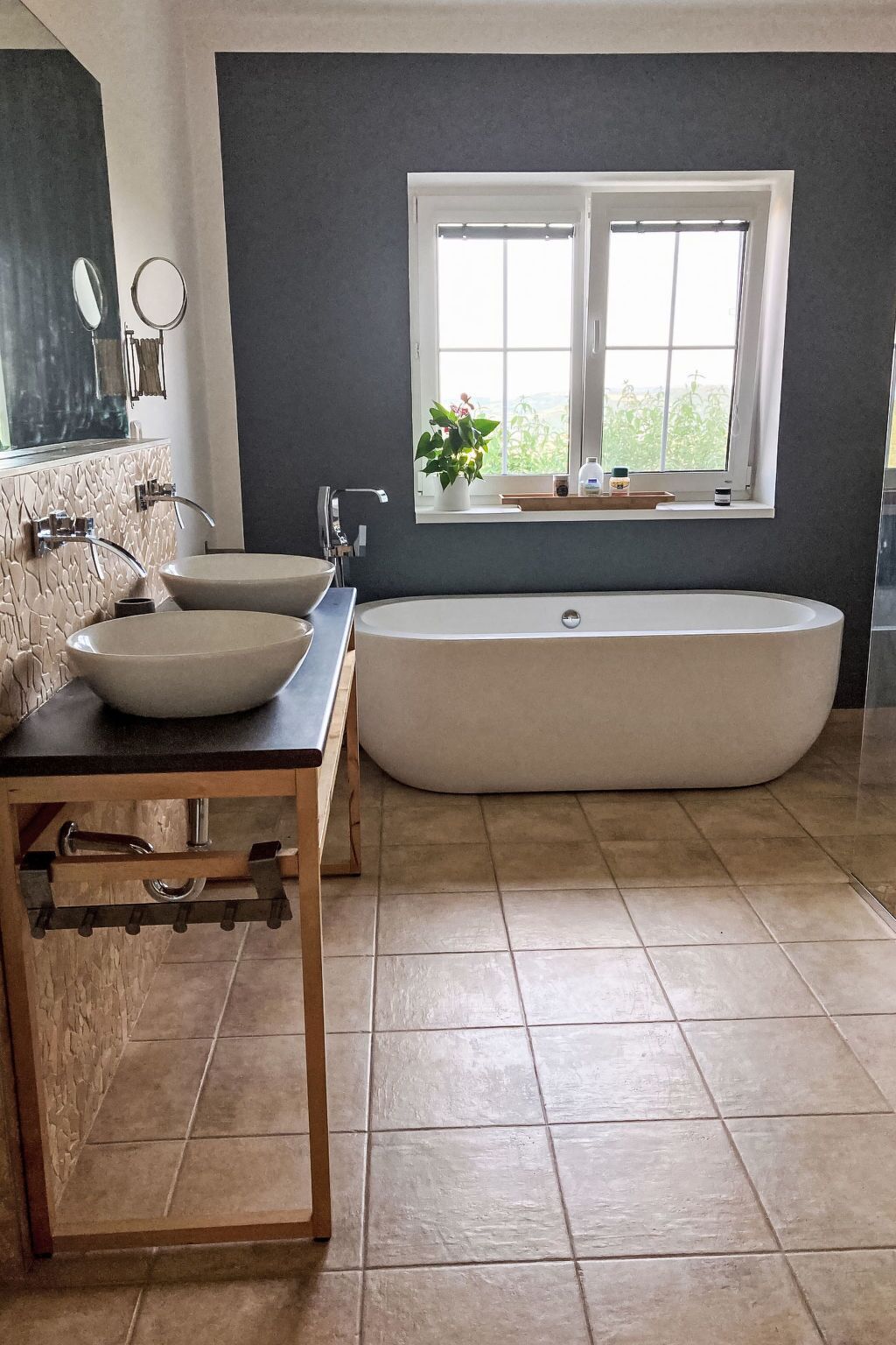 A modern bathroom with a freestanding bathtub, two vessel sinks, a wooden vanity, and a window with a view.