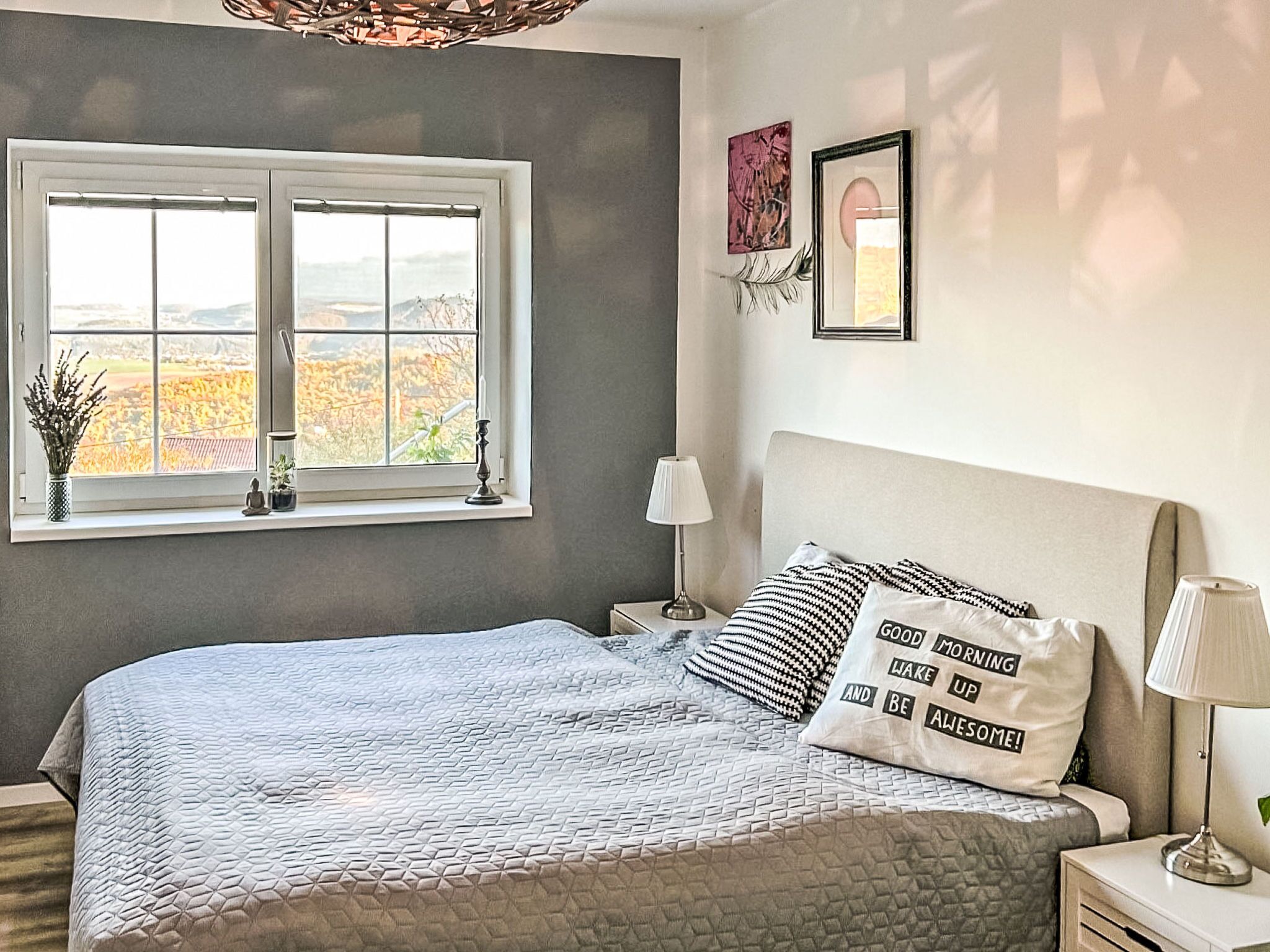 Un dormitorio con cama, ventana, mesita de noche, lámpara y una pared con dos cuadros enmarcados.