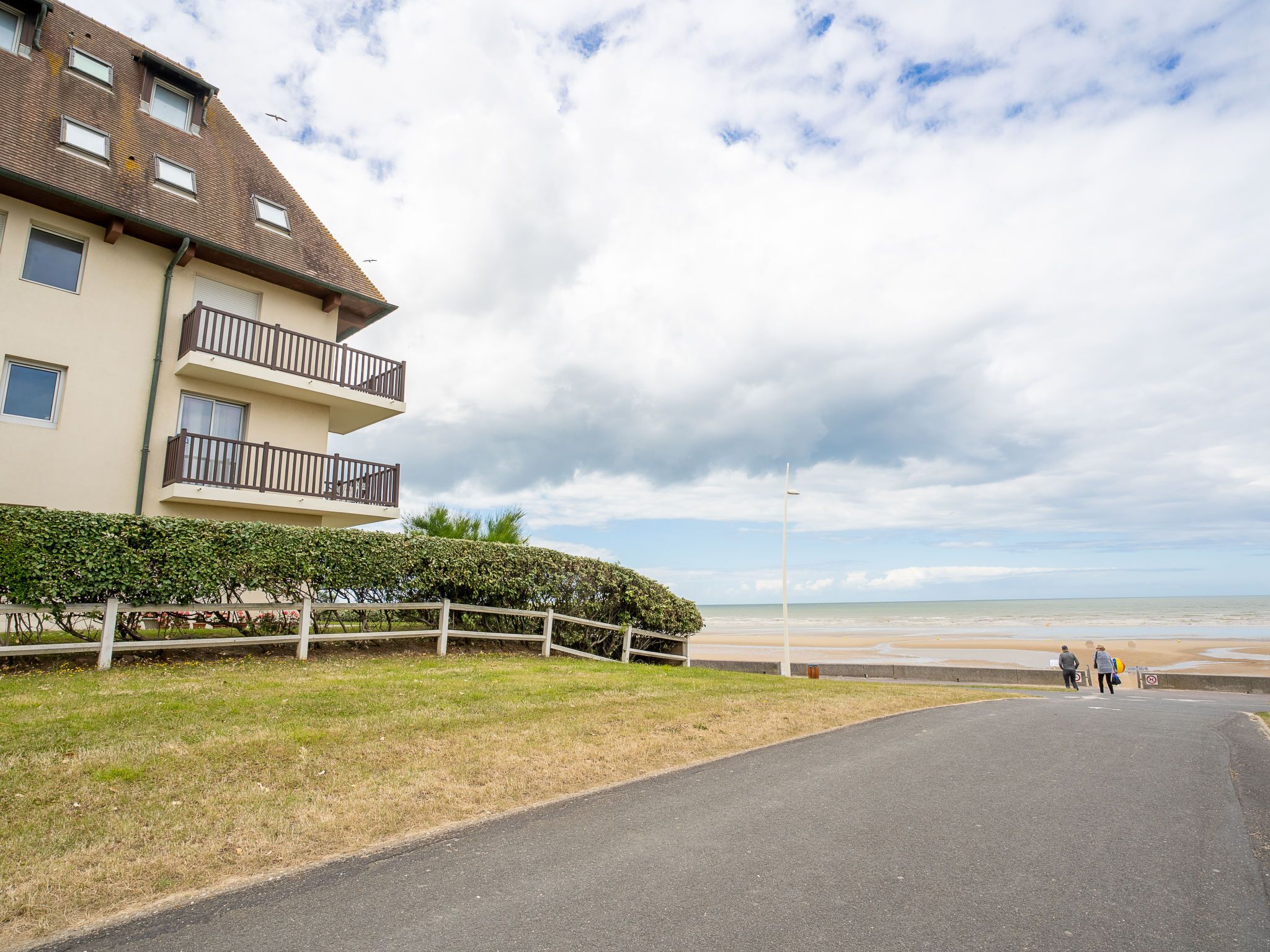 A multi-story building with balconies, a paved road, and a beach in the background.