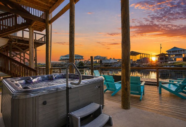 A hot tub on a wooden deck with chairs, overlooking a marina with houses.