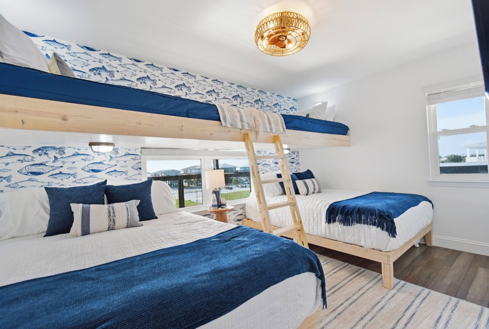 Une chambre avec un lit superposé et une échelle, deux lits avec une literie bleue, un lustre et une fenêtre donnant sur un beau paysage.