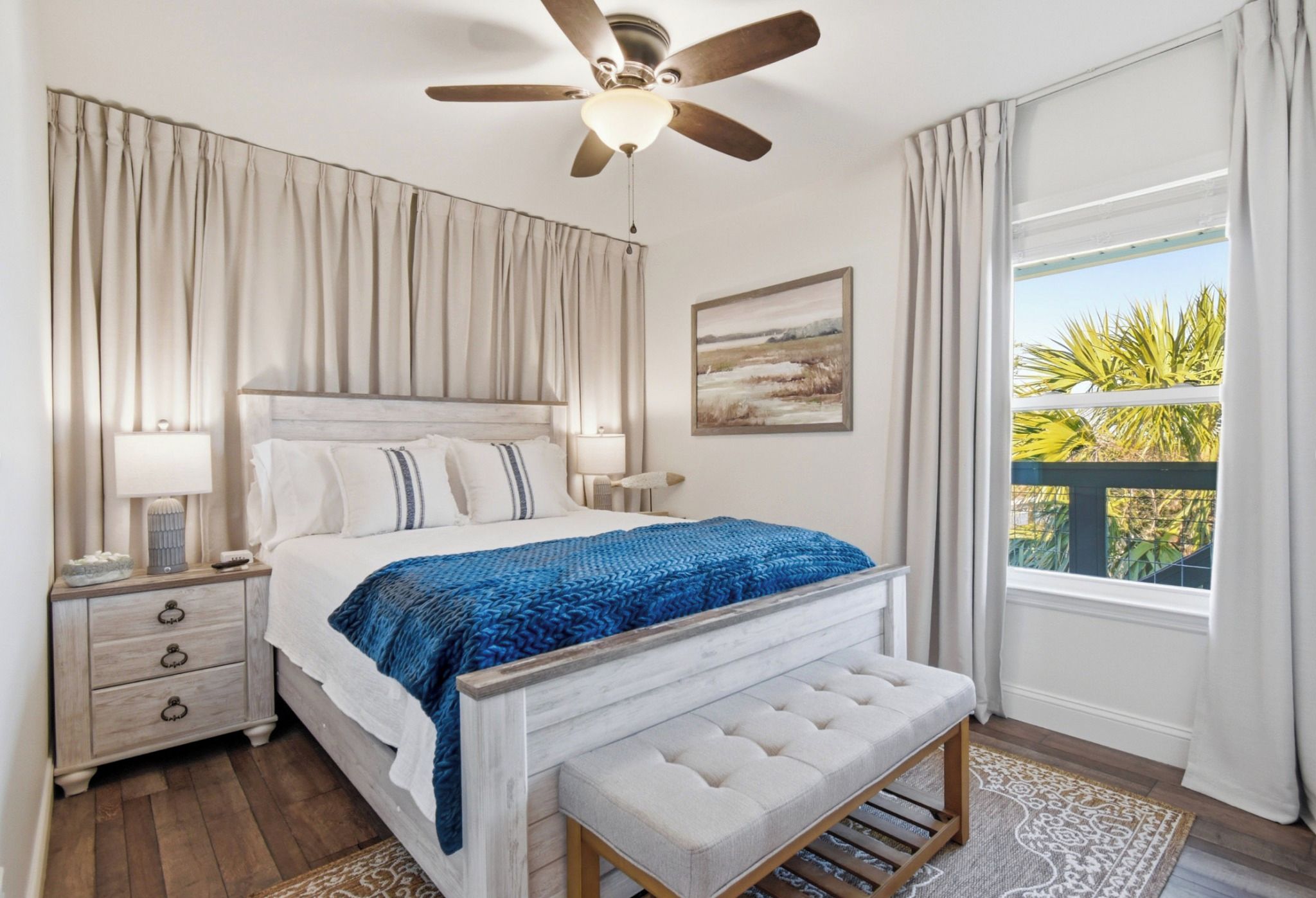 Une chambre à coucher avec un lit, des tables de chevet, un ventilateur de plafond, une fenêtre avec des rideaux et une vue sur la verdure à l’extérieur.