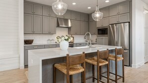 Cocina moderna con armarios grises, electrodomésticos de acero inoxidable, una isla blanca con taburetes de bar e iluminación colgante.