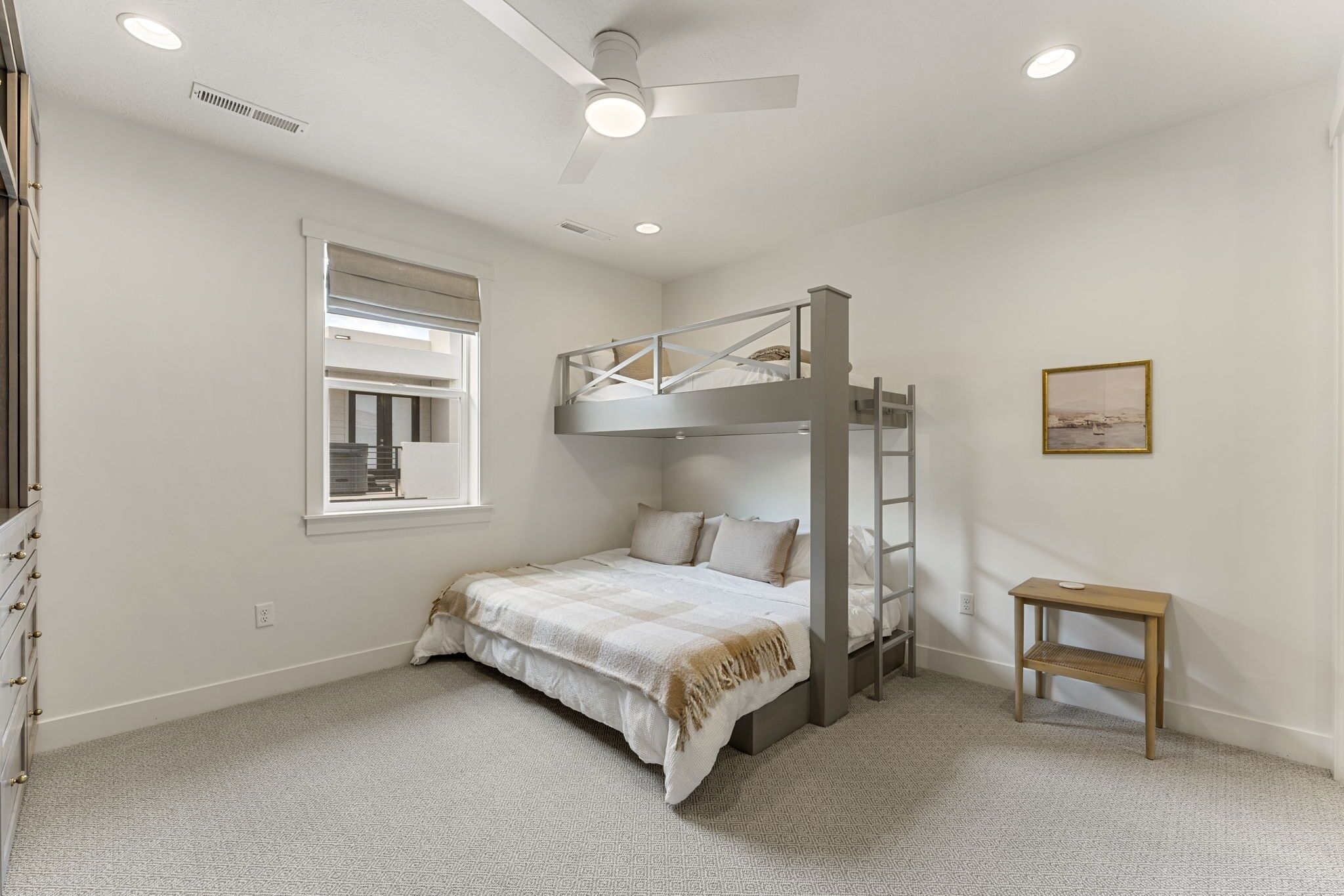 Quarto com beliche, ventilador de teto, janela com persianas, mesa de cabeceira e um quadro na parede.