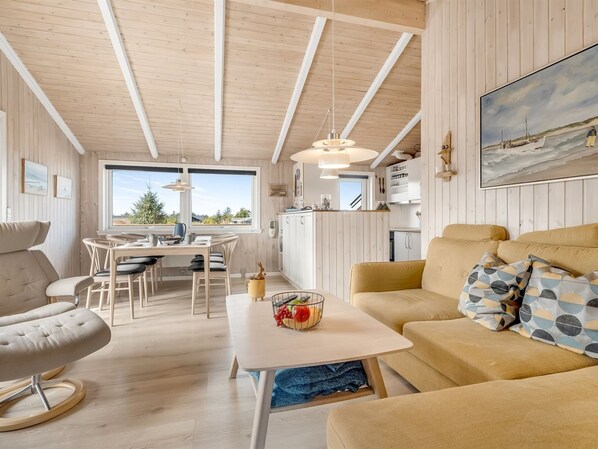 A cozy room with a wooden ceiling, a dining area with a table and chairs, a sofa with patterned cushions, and a painting on the wall.