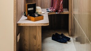 Um quarto pequeno revestido em madeira, com um banco, sapateira e cabideiro com várias peças de roupa.