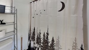 A bathroom with a shower curtain featuring a forest and moon design, a metal shelf with a basket, and a toilet paper roll. - Buck Deer Cabinette: Studio + Mountain Views! (Alto)