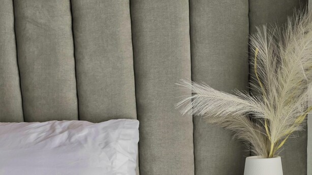 Una cama con un almohadón blanco y un florero con plantas secas sobre una mesita de noche.