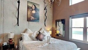 A bedroom with a bed, a nightstand, a lamp, a painting of the Mona Lisa, a chandelier, and a window. - Ocean Grove PH 1101 | Stunning Penthouse & Gulf Views! (Galveston)