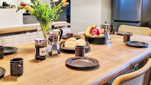 Une table à manger dressée avec des assiettes, une cafetière, un panier de fruits et un vase rempli de fleurs.
