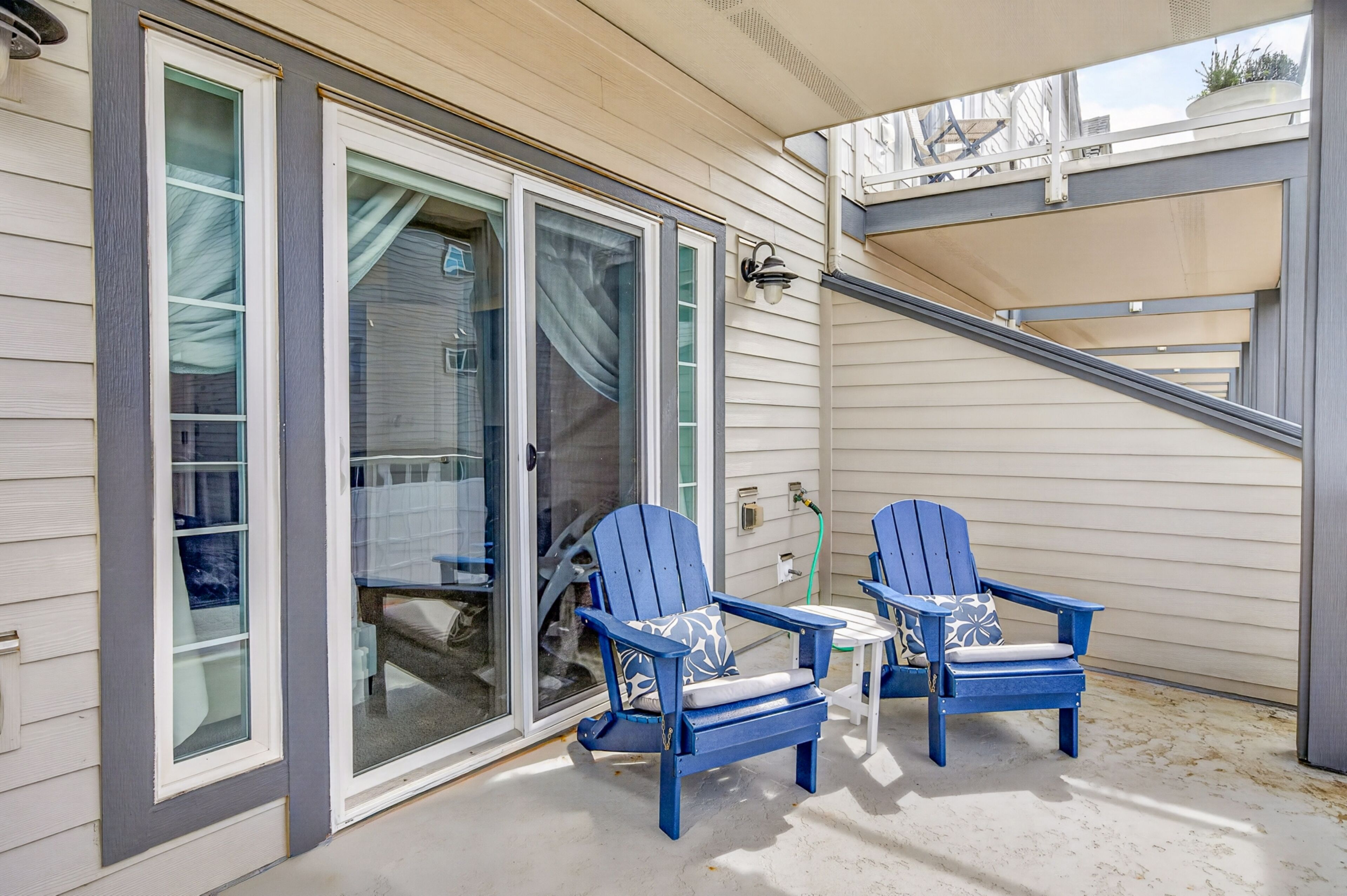 A covered outdoor area with two blue Adirondack chairs, a sliding glass door, and a small table.