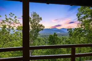 Blick durch ein Fenster mit Holzrahmen auf eine bewaldete Landschaft mit Bergen in der Ferne und einem klaren Himmel.