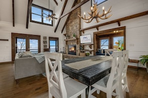 A spacious living area with a fireplace, a dining table with chairs, and a large window overlooking a body of water. - Views in the Blues: 3-Story Asheville Log Cabin with Hot Tub & Fire Pit (Arden)