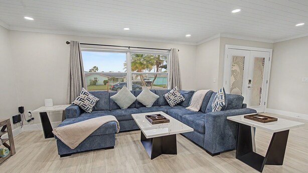 A modern living room with a sectional sofa, two coffee tables, and a view of a residential area. - Canal Front Getaway | Pool, Dock, Game Room + Screened Porch (Port Richey)