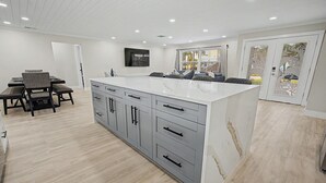 A modern kitchen with a large island, grey cabinets, and a dining area with chairs and a table. - Canal Front Getaway | Pool, Dock, Game Room + Screened Porch (Port Richey)
