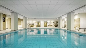 Una amplia piscina interior con suelo de baldosas, rodeada de paredes con una combinación de ladrillo y paneles blancos, y puertas que dan paso a otras zonas.