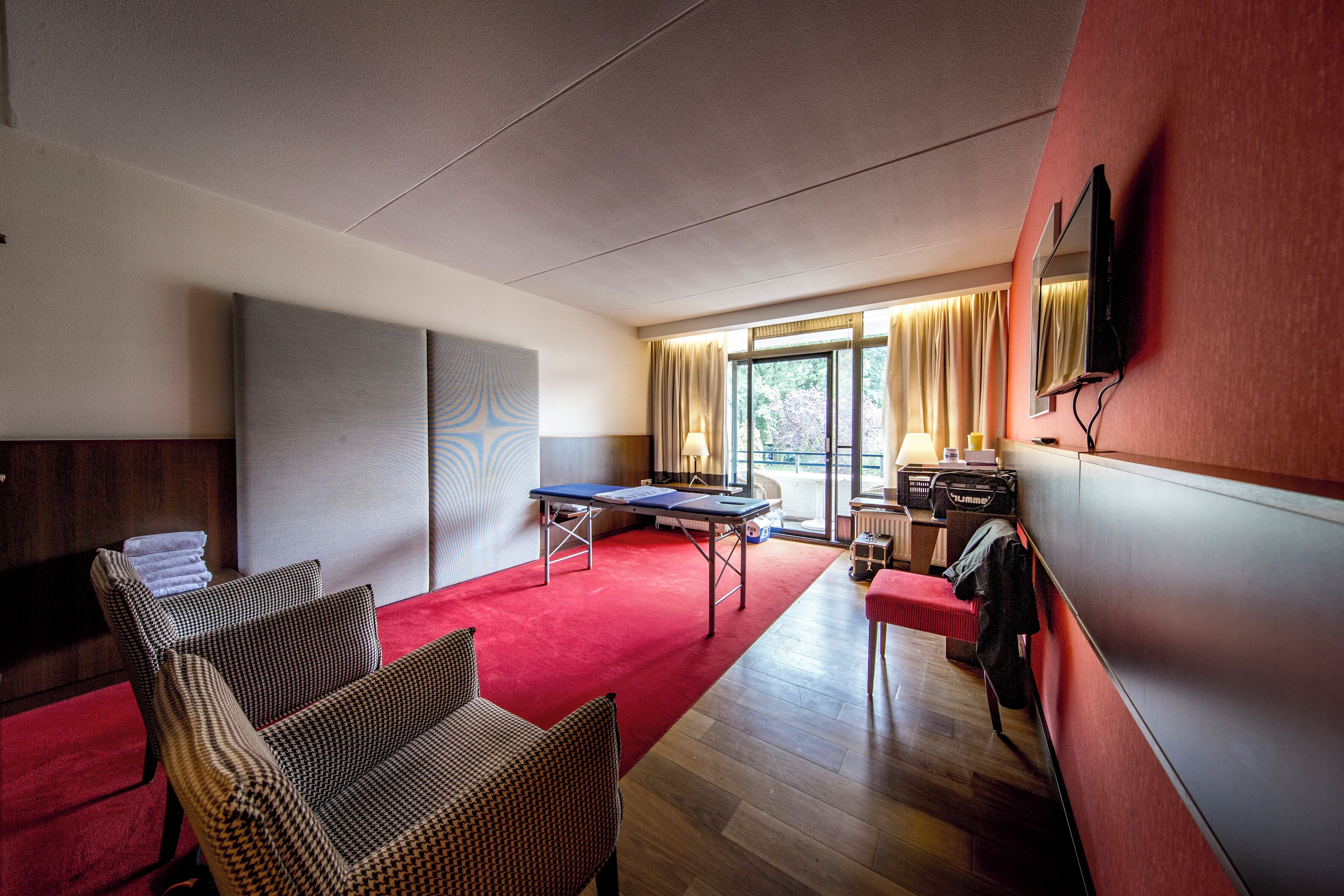 Habitación de hotel con alfombra roja, mesa de billar, dos sillas, un sofá, televisor y un ventanal grande con cortinas.
