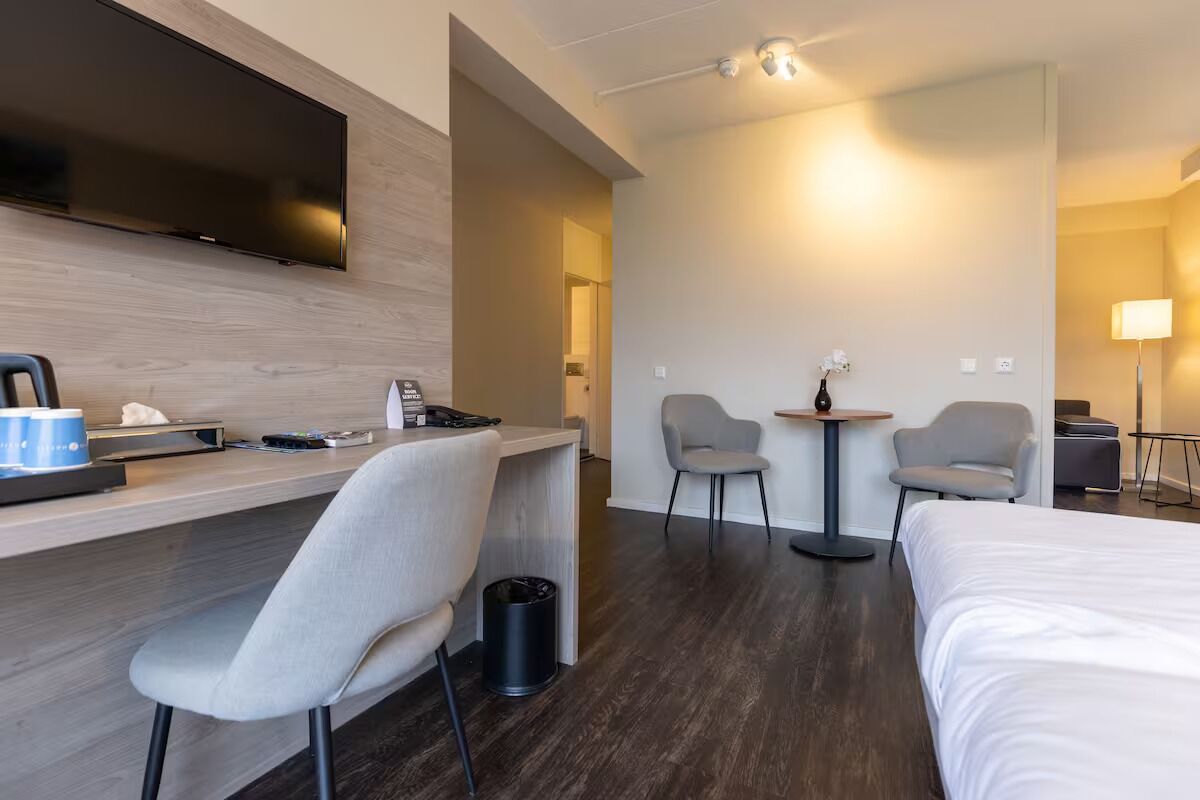 Una habitación de hotel con una cama, un escritorio con una silla, un televisor en la pared y dos sillones alrededor de una mesita.