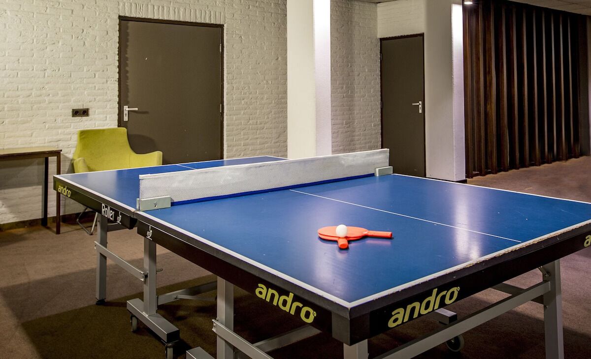 Un tavolo da ping pong con una racchetta arancione, una sedia gialla e un muro di mattoni bianchi.