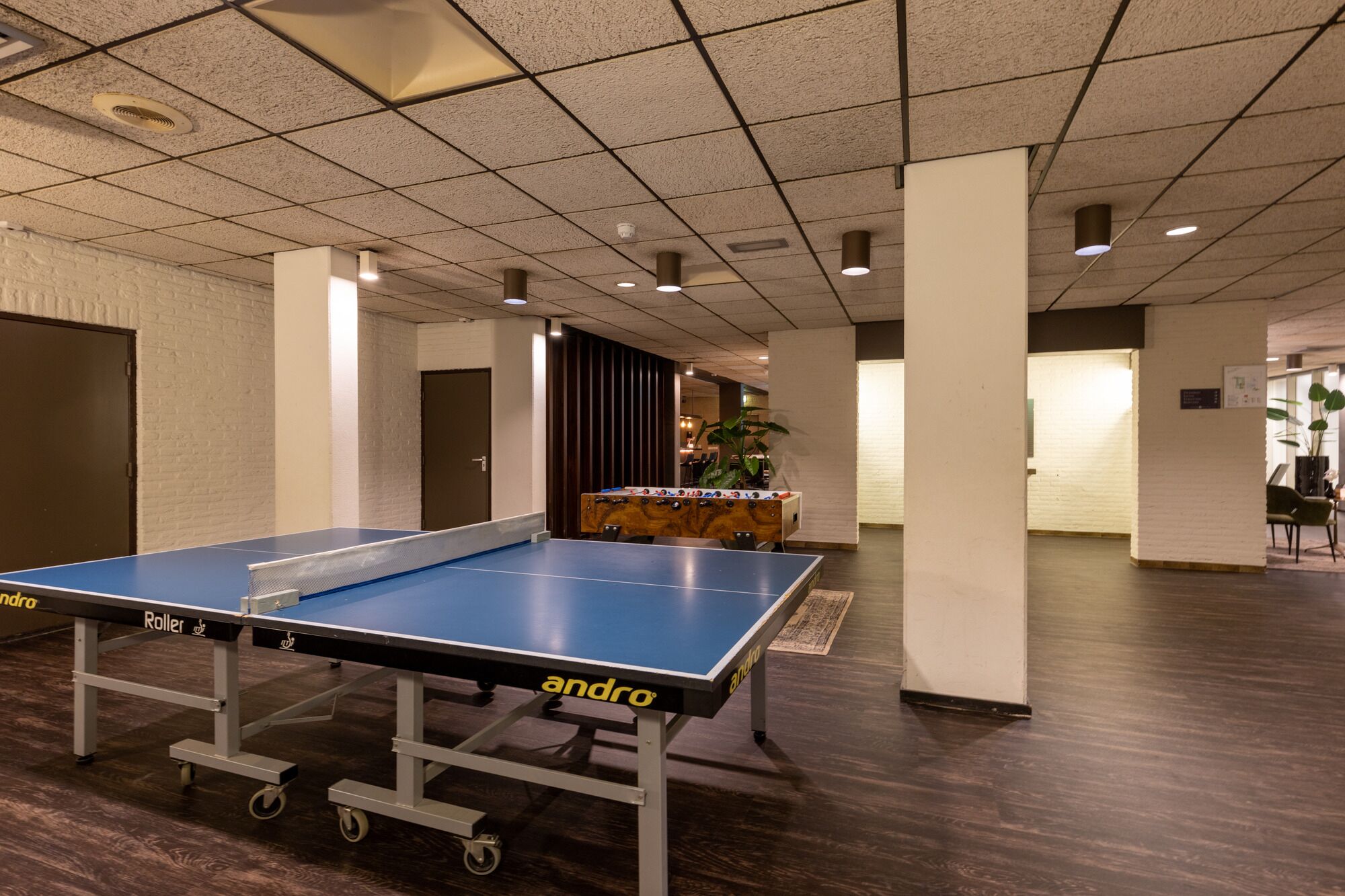 Ett bordtennisbord med hjul, ett fotbollsspel och en krukväxt i ett rum med ett kaklat tak och trägolv.