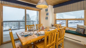Ett matbord i trä, dukat med tallrikar, glas och bestick, omgivet av stolar, med utsikt över ett snötäckt landskap genom stora fönster.