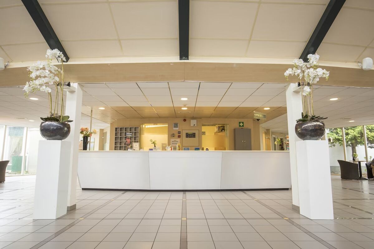 Een receptie met een wit receptiebalie, twee potplanten met witte bloemen, een betegelde vloer en een plafond met inbouwverlichting.