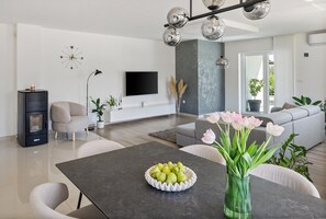 A modern living room with a flat-screen TV, a fireplace, a dining table with a fruit bowl, and a sofa. - Villa Green Escape: Your Serene Family Getaway Awaits (Grubine)