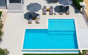 Ein rechteckiger Swimmingpool mit einer gefliesten Liegefläche darum. Liegestühle und Sonnenschirme sind rund um den Pool angeordnet.