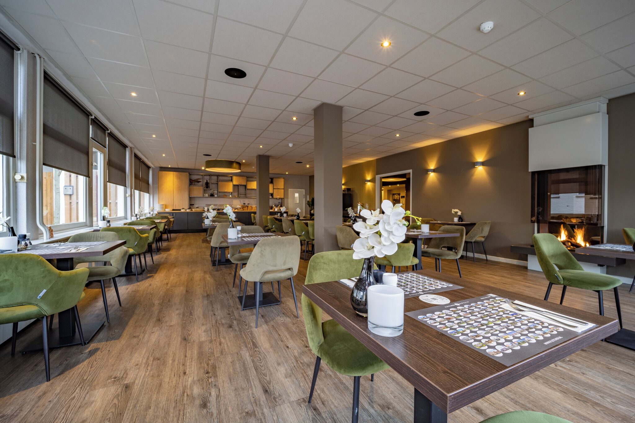 Een ruime hotel eetzaal met meerdere tafels en stoelen, een centraal bloemstuk, en een uitzicht op de keuken op de achtergrond.