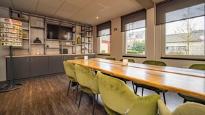 Een vergaderruimte met een lange tafel, groene stoelen, een boekenkast en een raam met uitzicht op gebouwen.