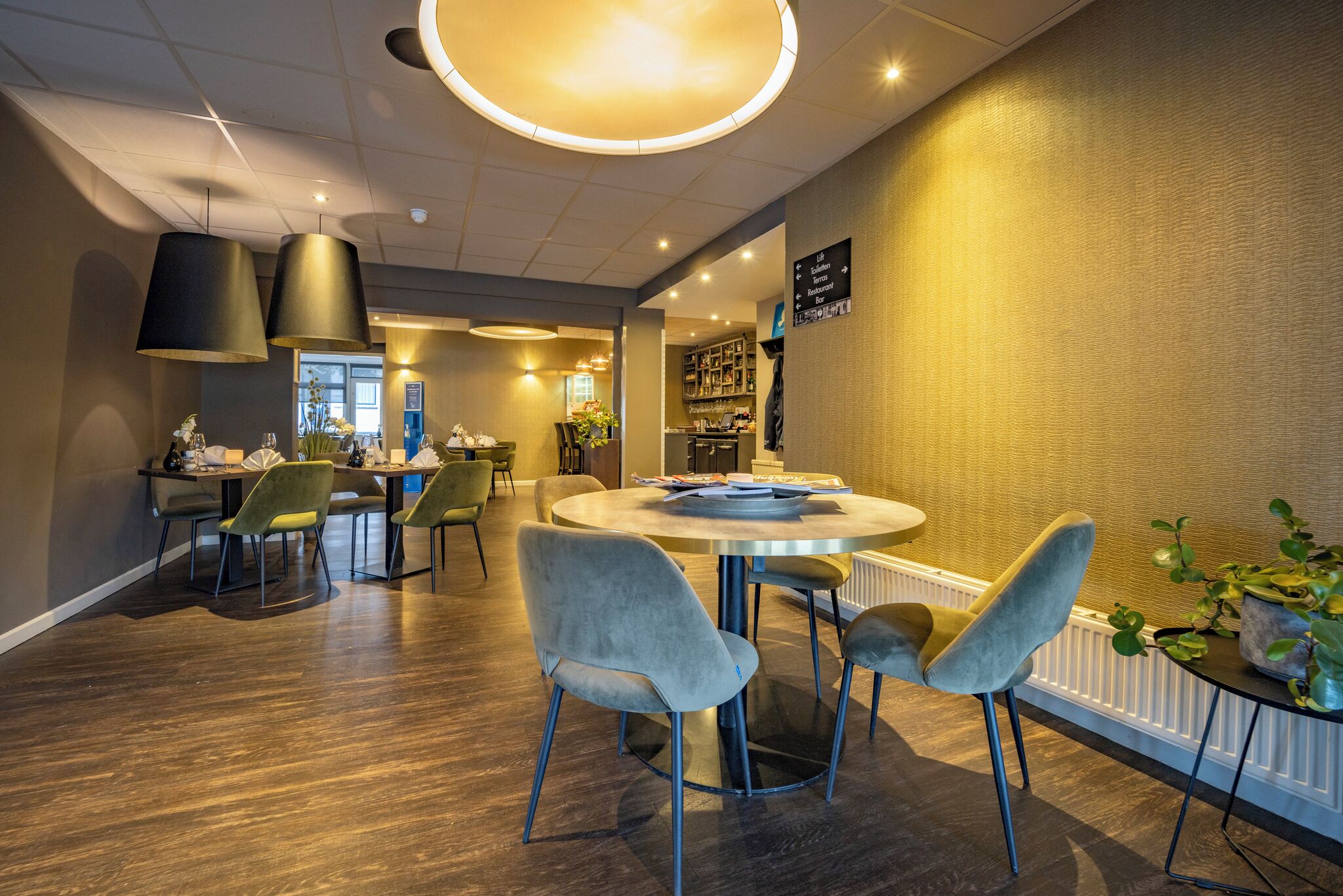 Een moderne eetruimte met een ronde tafel, gestoffeerde grijze stoelen, hanglampen en een zichtbare keuken op de achtergrond.