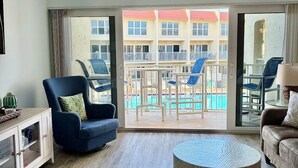 Quarto de hotel com varanda, ventilador de teto, sofá, mesa de centro e vista para uma piscina e edifícios.