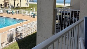 Uma piscina com vista para a praia, um edifício com palmeiras e uma varanda com guarda-corpo.