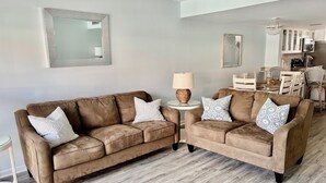 Uma sala de estar com dois sofás marrons, uma mesa de apoio redonda com abajur, e um espaço de jantar com mobiliário branco.