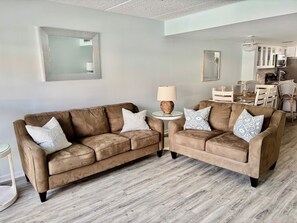 A living room with two brown sofas, a round side table with a lamp, and a dining area with white furniture. - 2 BR/2.5 Bath Beach Condo in St. Augustine- walking distance to food & fun (St. Augustine)