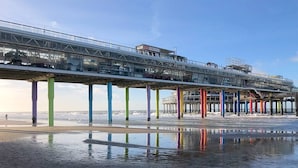 Eine Pier mit einer Glasgalerie, gestützt von bunten Säulen, die sich über einen feuchten Strand erstreckt.