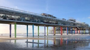 Eine Pier mit einer Glasgalerie, gestützt von bunten Säulen, die sich über einen feuchten Strand erstreckt.