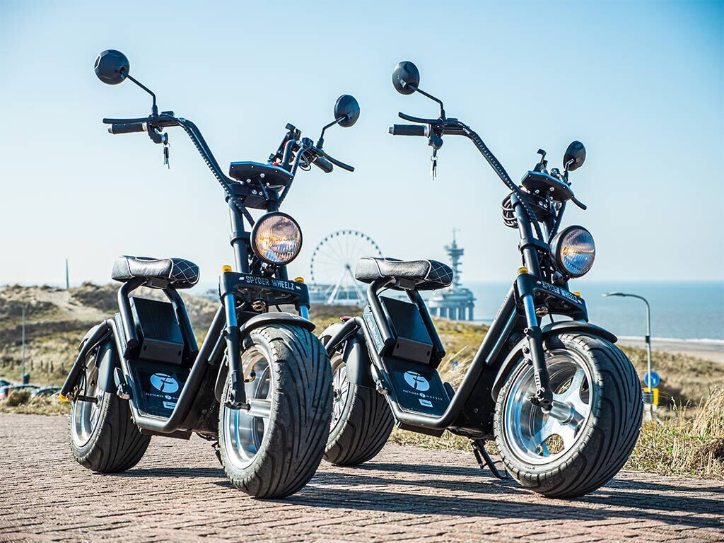 Two electric scooters parked on a paved surface with a clear sky and a distant Ferris wheel in the background.