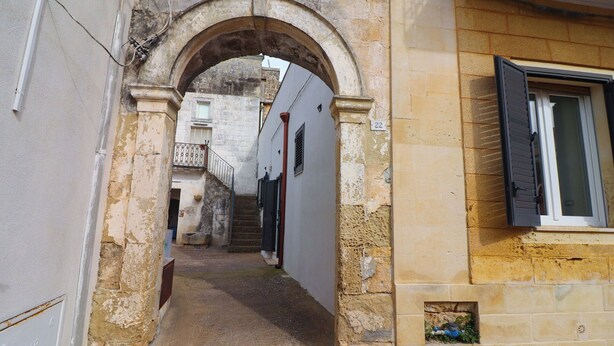 Une ruelle étroite, surmontée d’une arche en pierre, mène à un bâtiment doté d’une fenêtre et d’une porte.