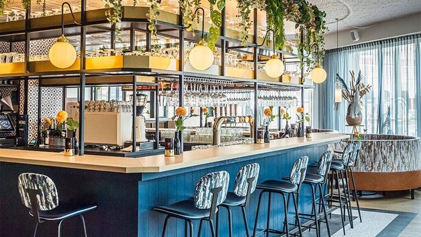 Eine Bar mit einer Theke, Barhocker, hängenden Pflanzen und Blick auf die Stadt durch große Fenster.