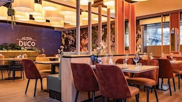 Interior de restaurante con mesas de madera, sillas de terciopelo rojo, plantas colgantes e iluminación cálida.