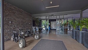 Entrada de un edificio moderno con un muro de piedra, dos scooters eléctricos y un puerta de cristal que da al interior.