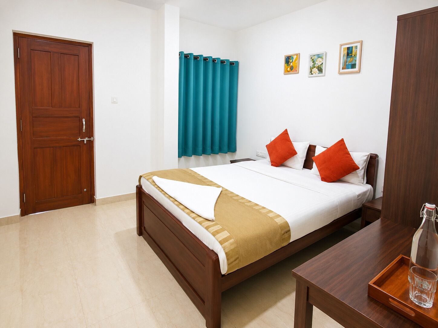 Una habitación de hotel con puerta de madera, cama con ropa blanca y almohadas anaranjadas, cortina color turquesa y una mesita con un vaso y una botella.