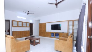 Una sala de estar con dos sofás, una mesa de centro, armarios de madera, un televisor de pantalla plana, ventiladores de techo y una puerta con cortina.