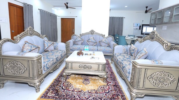 A room with a sofa set, a coffee table, a patterned rug, and a ceiling fan. - SBHS HOMESTAY-SREE ANJANADHRI (Tirupati)
