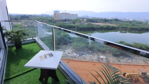 Un balcón con barandilla de vidrio, una mesita con una revista y vista a un río y edificios al fondo.