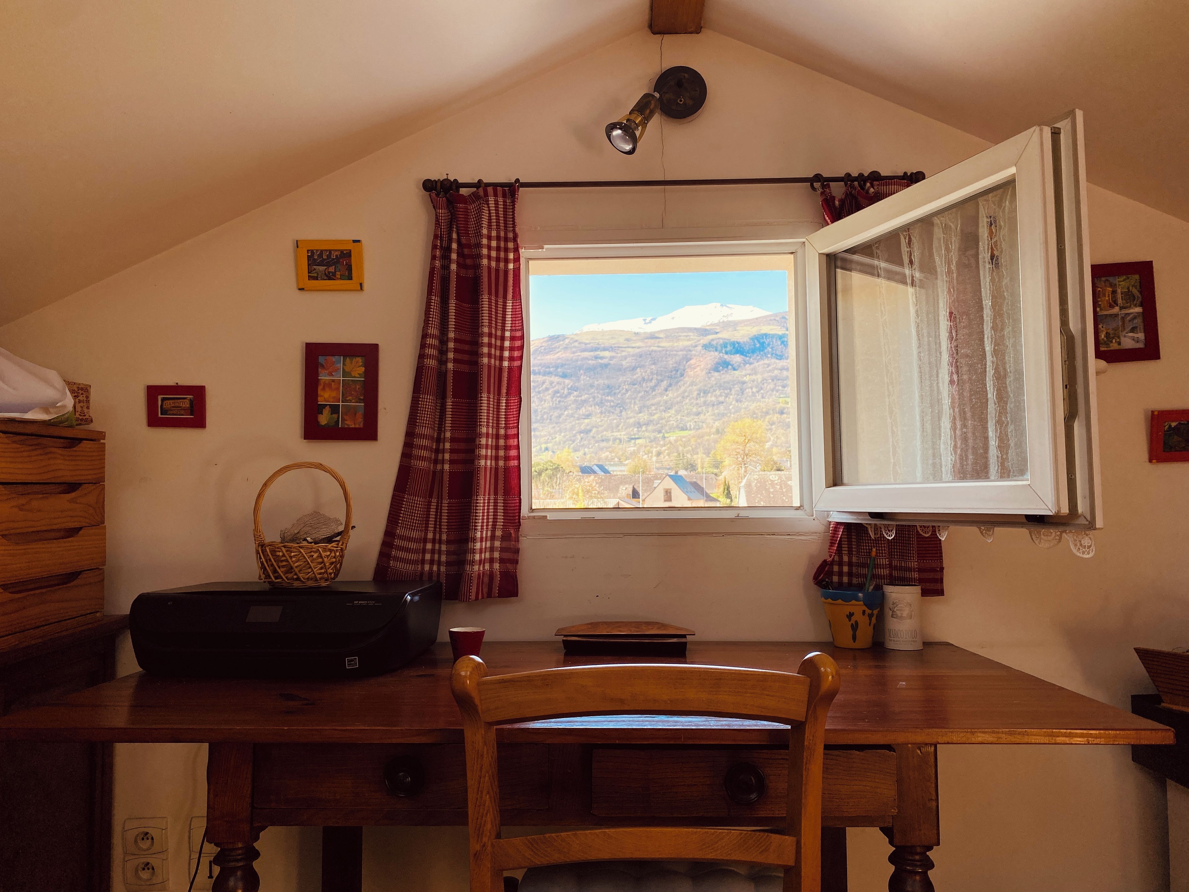 Quarto com escrivaninha, cadeira, janela com cortinas e vista para uma paisagem montanhosa.