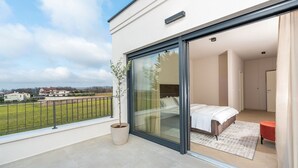Une chambre moderne dotée d’une grande porte-fenêtre coulissante donnant sur un balcon, d’un lit avec une literie blanche et d’une petite table rouge.