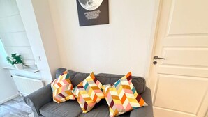 Un sofá gris con almohadones de diseño colorido en patrón de rombos, un radiador blanco, un cuadro en la pared y una puerta blanca.